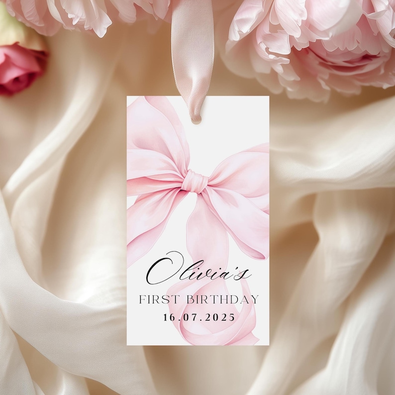 May include: A white gift tag with a watercolor pink bow design and the text "Olivia's First Birthday 16.07.2025". The tag is tied with a pink ribbon and set against a backdrop of soft fabric and pink flowers.