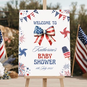 May include: A white sign on a wooden easel with patriotic decorations. The sign reads "WELCOME TO Katherine's BABY SHOWER 4th July 2025" with a bow in the colors of the American flag. Red, white, and blue accents.