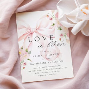 Puede incluir: Una invitación de despedida de soltera rosa y blanca con una ilustración de acuarela de un lazo rosa y flores. El texto dice "Love in Bloom" e incluye el nombre de la novia, la fecha, la hora y el lugar.