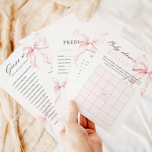 Puede incluir: Una mano sosteniendo un montón de tarjetas de juegos para baby shower. Las tarjetas presentan lazos rosas de acuarela y texto, incluyendo "Guess us", "Predictions" y "Baby shower BINGO". El fondo es de un color suave y neutro.