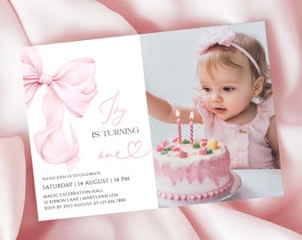 May include: A pink birthday invitation with a watercolour bow and the text "Joy is turning one." The invitation features a photo of a baby girl with a pink cake and candles. The invitation details include the date, time, and location.