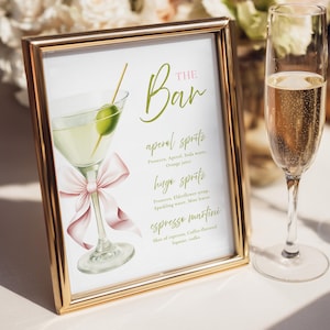 May include: A gold-framed sign with a cocktail illustration and drink menu. The sign features a martini with an olive, a pink bow, and the words "THE Bar" in green script. The menu lists Aperol Spritz, Hugo Spritz, and Espresso Martini.