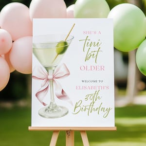 May include: A white sign on an easel with a martini glass illustration and text that reads "She's a tini bit OLDER, Welcome to Elisabeth's 30th Birthday." Pink and green balloons are in the background.