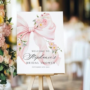 Puede incluir: Un cartel blanco con un lazo rosa y un diseño floral. El cartel dice "Bienvenido al Bridal Shower de Stephanie, 16 de noviembre de 2023".