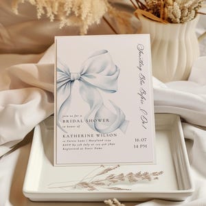 May include: A blue and white bridal shower invitation with a watercolor illustration of a bow. The invitation reads "Something Blue Before Do!" and includes the text "Join us for a Bridal Shower in honor of Katherine Wilson" and the date and time of the event.