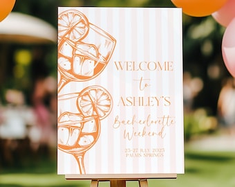 May include: A welcome sign on an easel with a cocktail illustration. The sign reads "Welcome to Ashley's Bachelorette Weekend" with the dates July 25-27, 2023, and "Palais Springs" below. The background has pink and white stripes.
