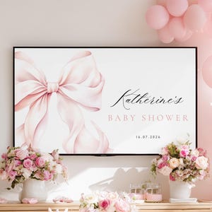 May include: A baby shower sign with a large pink bow illustration and the text "Katherine's BABY SHOWER 16.07.2026". The sign is framed in black and surrounded by pink balloons and floral arrangements.