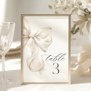May include: A gold-framed table number card with a white satin bow and the text "table 3".