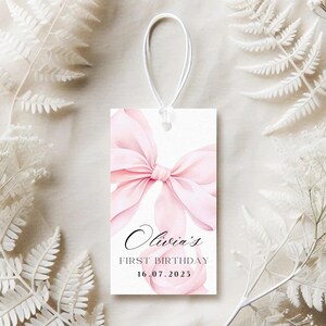 May include: A rectangular gift tag with a watercolor pink bow design. The tag reads "Olivia's FIRST BIRTHDAY 16.07.2025" in elegant script and serif fonts. A white string is attached at the top for hanging. The background is white.