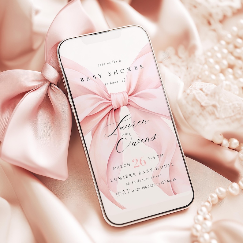 May include: A smartphone displays a baby shower invitation with a large pink bow graphic. The invitation text includes the name "Lauren Owens", date, time, and RSVP information. The phone is set against a backdrop of pink fabric and a matching bow.