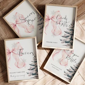 Op de afbeelding: Vier ingelijste borden met een lichte houten afwerking. Elk bord heeft een aquarelontwerp van een roze strik, sneeuwvlokken en een dennenboom. Tekst op de borden is onder meer "S'mores Bar", "Cards & Gifts", "Favors" en "Diaper Raffle".