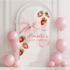 May include: A baby shower backdrop featuring a large pink bow and watercolor strawberries. The text "Amelie's BABY SHOWER" is displayed in a delicate script. Pink balloons of various sizes are arranged around the backdrop.