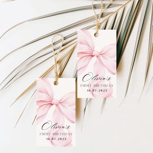 May include: Two rectangular gift tags with a watercolor pink bow design and the text "Olivia's First Birthday 16.07.2025". Each tag is suspended by a natural twine string.