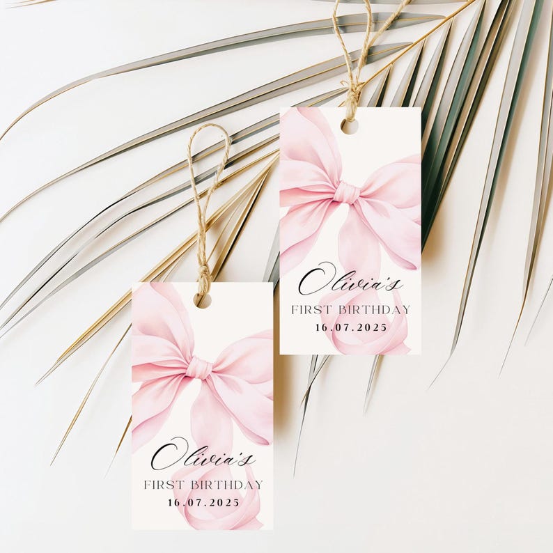 May include: Two rectangular gift tags with a watercolor pink bow design and the text "Olivia's First Birthday 16.07.2025". Each tag is suspended by a natural twine string.