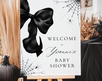 May include: A white sign with a black bow and spiderwebs, welcoming guests to "Yvonne's Baby Shower." The sign is displayed on a wooden easel, with pumpkins and decorative elements in the background, creating a Halloween-themed baby shower atmosphere.