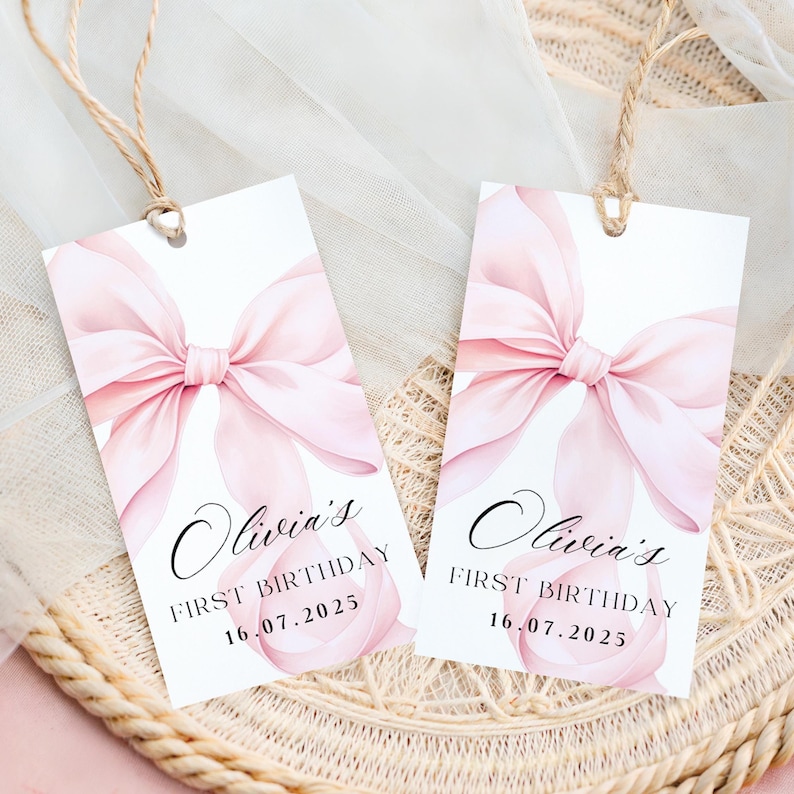 May include: Two white gift tags with a watercolor pink bow design. The tags read "Olivia's First Birthday 16.07.2025" in elegant script. Each tag has a natural twine string for hanging.