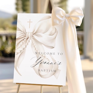 Puede incluir: Un letrero de bienvenida blanco para el bautizo de Yvonne, con un gran lazo y detalles de perlas. El letrero incluye el texto "Welcome to Yvonne's Baptism" y se muestra en un caballete dorado. Un lazo a juego está adjunto al lado.