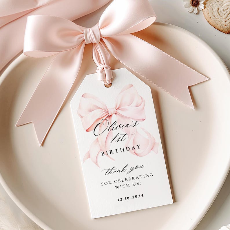 Pode incluir: Uma etiqueta branca com uma ilustra&ccedil;&atilde;o de la&ccedil;o rosa e o texto "Olivia's 1st Birthday" e "Thank you for celebrating with us! 12.10.2024". A etiqueta est&aacute; amarrada com uma fita de cetim rosa.