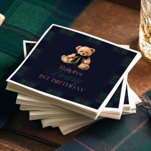 May include: A pile of navy blue square cards featuring a teddy bear illustration and a plaid border. The text reads "Ralph's Beary 1st Birthday". The cards are stacked on a wooden surface, with a tartan fabric in the background.