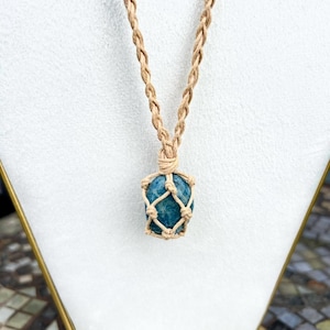 May include: A blue gemstone pendant necklace. The gemstone is encased in a beige macrame net and suspended from a braided beige cord. The necklace is displayed on a white stand.