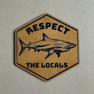 以下が含まれることがあります： 黒いサメのシルエットと「RESPECT THE LOCALS」というテキストが書かれた茶色の六角形のステッカーです。
