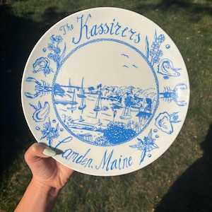 May include: A white ceramic plate featuring a blue illustration of a harbor with sailboats, surrounded by a border of oysters, lobsters, and floral motifs. The text reads The Kassirers at the top and Camden, Maine at the bottom in blue script.