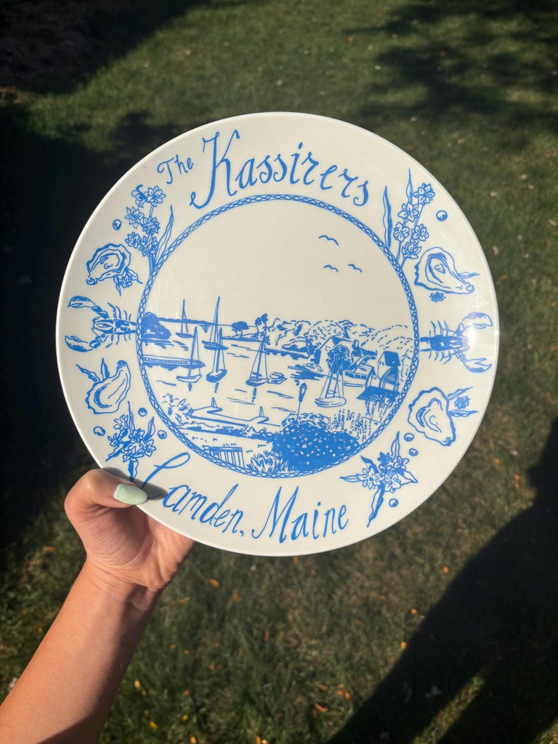 May include: A white ceramic plate featuring a blue illustration of a harbor with sailboats, surrounded by a border of oysters, lobsters, and floral motifs. The text reads The Kassirers at the top and Camden, Maine at the bottom in blue script.