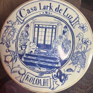 May include: A round white ceramic plate with a gold rim featuring a blue ink illustration of a house entrance. The text Casa Lark de Luz is at the top and KOLOA, HI is at the bottom. The design includes a sea turtle, a bird, and tropical plants.