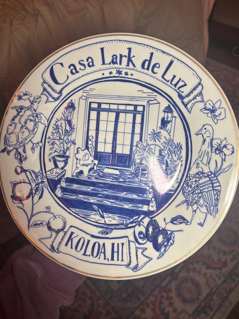 May include: A round white ceramic plate with a gold rim featuring a blue ink illustration of a house entrance. The text Casa Lark de Luz is at the top and KOLOA, HI is at the bottom. The design includes a sea turtle, a bird, and tropical plants.