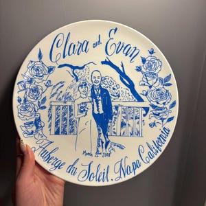 May include: A round white ceramic plate featuring a blue line drawing of a couple standing on a balcony with rose borders. The text reads Clara and Evan, March 22nd 2008, and Auberge du Soleil, Napa California in a decorative script font.