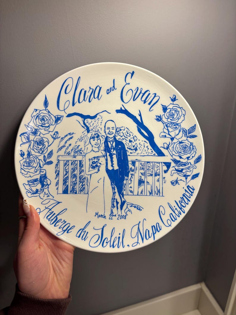 May include: A round white ceramic plate featuring a blue line drawing of a couple standing on a balcony with rose borders. The text reads Clara and Evan, March 22nd 2008, and Auberge du Soleil, Napa California in a decorative script font.