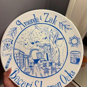May include: A round white ceramic plate featuring blue line art illustrations. The center depicts an outdoor patio scene with tables and trees. Surrounding the image are a dog, a cigar, a sun, and a glass. Text reads Amanda & Zak and Bacari Sherman Oaks.