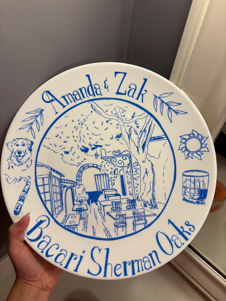 May include: A round white ceramic plate featuring blue line art illustrations. The center depicts an outdoor patio scene with tables and trees. Surrounding the image are a dog, a cigar, a sun, and a glass. Text reads Amanda & Zak and Bacari Sherman Oaks.
