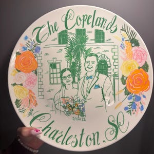 May include: A round white ceramic plate featuring a green line drawing of a couple in front of a building with a palm tree. The text reads The Copelands, 9/26/22, and Charleston, SC. The design is framed by colorful painted flowers in orange, yellow, and pink.