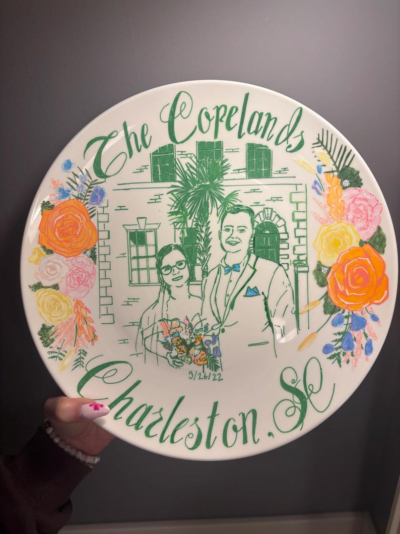 May include: A round white ceramic plate featuring a green line drawing of a couple in front of a building with a palm tree. The text reads The Copelands, 9/26/22, and Charleston, SC. The design is framed by colorful painted flowers in orange, yellow, and pink.