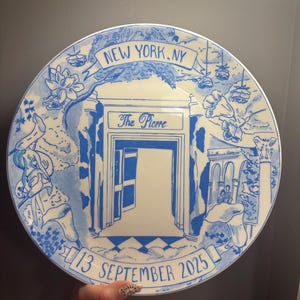 May include: A round white ceramic plate featuring blue hand-painted illustrations of The Pierre hotel entrance in New York, NY. The design includes floral accents, a banner reading NEW YORK, NY, and the date 13 SEPTEMBER 2025 at the bottom.