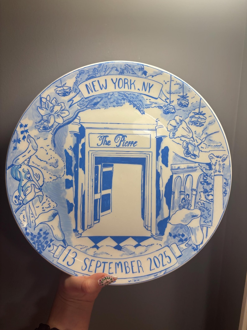 May include: A round white ceramic plate featuring blue hand-painted illustrations of The Pierre hotel entrance in New York, NY. The design includes floral accents, a banner reading NEW YORK, NY, and the date 13 SEPTEMBER 2025 at the bottom.