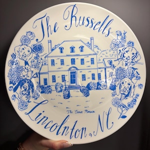 May include: A white ceramic plate featuring blue line art of a large house, dogs, and floral accents. The text reads The Russells, The Crowe Mansion, and Lincolnton, NC. The design is centered on the plate with a decorative script and illustration.