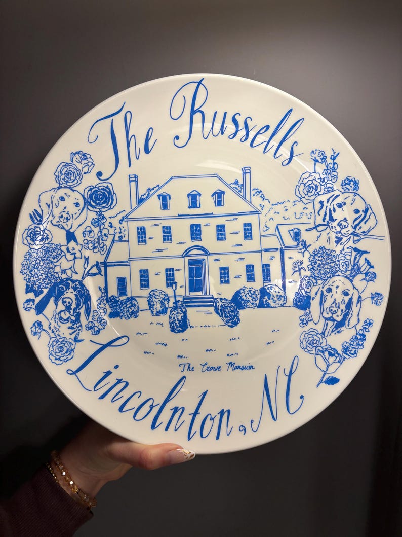 May include: A white ceramic plate featuring blue line art of a large house, dogs, and floral accents. The text reads The Russells, The Crowe Mansion, and Lincolnton, NC. The design is centered on the plate with a decorative script and illustration.