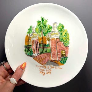 Op de afbeelding: Witte keramische plaat met een handgeschilderde afbeelding van een bakstenen ingang, een zwarte poort en bomen. De plaat bevat de tekst "University of South Carolina Hope 2026 Molly 2019". De plaat heeft een glanzende afwerking.
