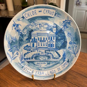 May include: A round white ceramic plate featuring a blue hand-painted illustration of Villa Erba on Lake Como. The text reads CHLOE and CYRUS at the top, and VILLA ERBA, LAGO DI COMO with the date August 16th, 2026 at the bottom, framed by olive branches.