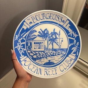 May include: A round white ceramic plate featuring a blue illustration of a coastal building with palm trees. The text reads The BOURGEOIS'S OCEAN REEF CLUB and the date 8-15-2015. The design includes floral accents and a decorative border.