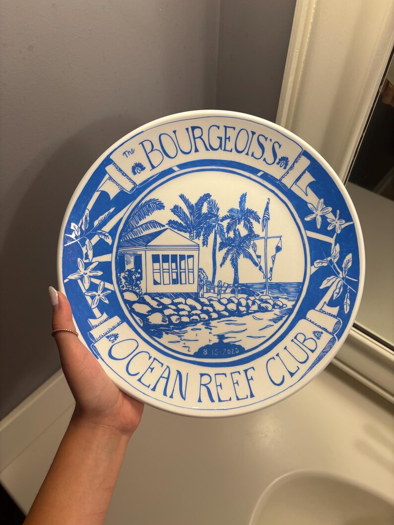 May include: A round white ceramic plate featuring a blue illustration of a coastal building with palm trees. The text reads The BOURGEOIS'S OCEAN REEF CLUB and the date 8-15-2015. The design includes floral accents and a decorative border.