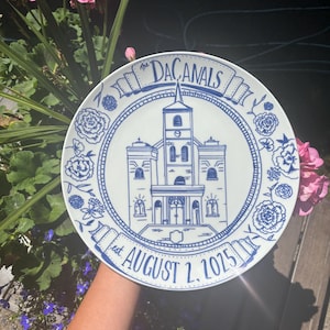 May include: A white ceramic plate featuring a blue line-art illustration of a church building in the center. The text "The DACANALS" is written in a banner at the top, and "est. AUGUST 2, 2025" is written in a banner at the bottom, surrounded by floral motifs.