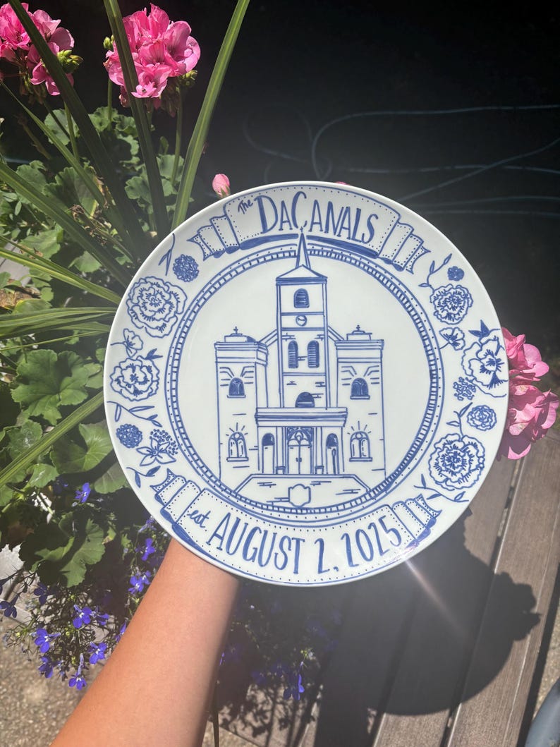 May include: A white ceramic plate featuring a blue line-art illustration of a church building in the center. The text "The DACANALS" is written in a banner at the top, and "est. AUGUST 2, 2025" is written in a banner at the bottom, surrounded by floral motifs.