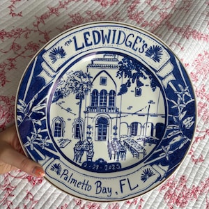 May include: A round ceramic plate featuring a blue and white illustration of a building with palm trees and outdoor dining tables. The text reads The LEDWIDGE'S, 2-28-2023, and Palmetto Bay, FL. The plate has a gold-toned rim and decorative floral borders.