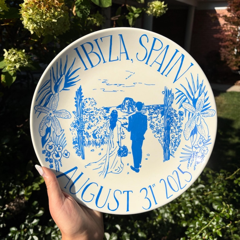 May include: A round white ceramic plate featuring a blue line art illustration of a couple walking toward a distant rock formation. The text reads IBIZA, SPAIN at the top and AUGUST 31ST 2025 at the bottom, framed by floral and palm leaf motifs.