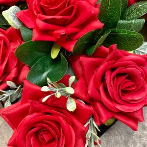 May include: Close-up of vibrant red roses with lush green leaves and delicate sprigs of gray-green foliage. The roses are in full bloom, showcasing their velvety petals. The arrangement is a classic floral design, perfect for various decorative purposes.