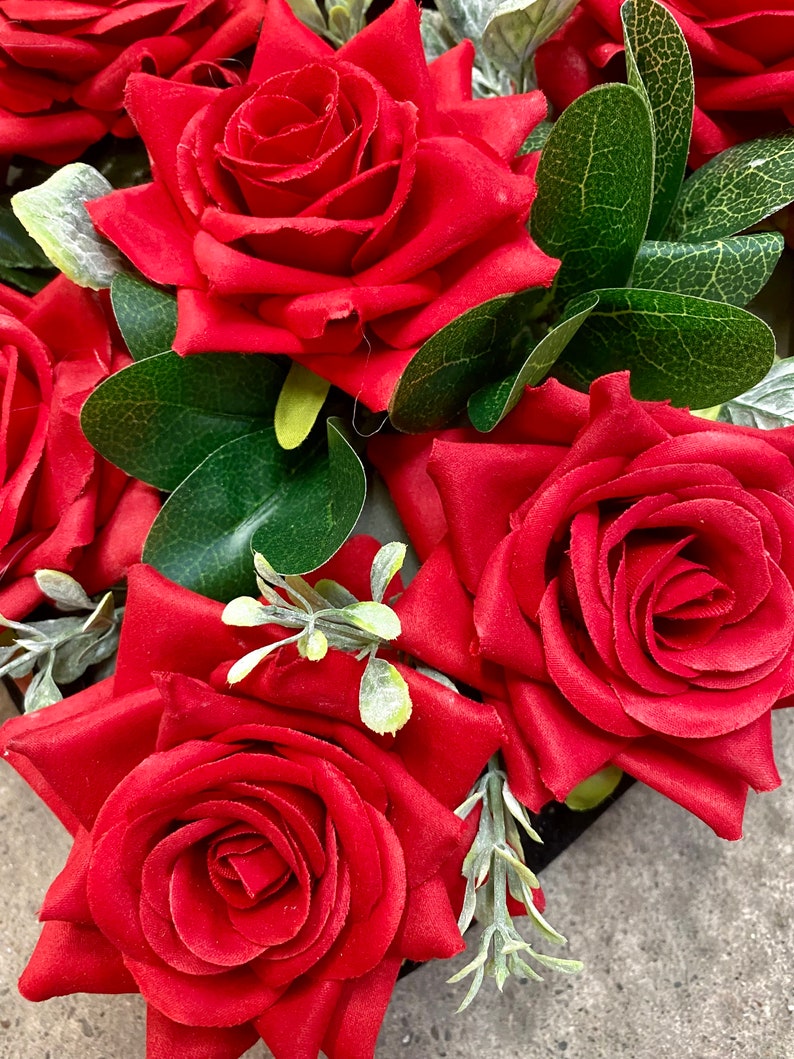 May include: Close-up of vibrant red roses with lush green leaves and delicate sprigs of gray-green foliage. The roses are in full bloom, showcasing their velvety petals. The arrangement is a classic floral design, perfect for various decorative purposes.