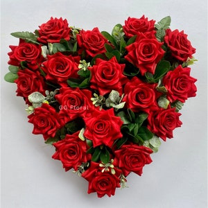 May include: A heart-shaped floral arrangement filled with vibrant red roses and green foliage. The roses are densely packed, creating a full and lush appearance. The arrangement includes the text "GG Floral".